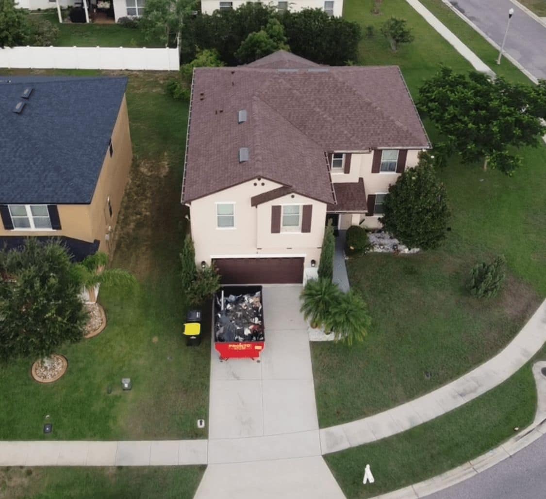 Drone view of a two-story home with a brown shingle roof, a red debris container in the driveway and a neighboring home with a dark blue-gray roof.