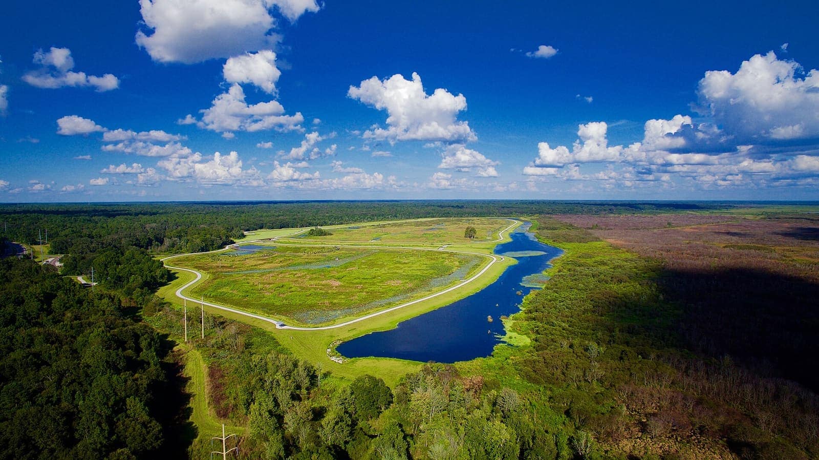 Aerial view of a green wetland park with a winding road, blue water channel and scattered clouds overhead.