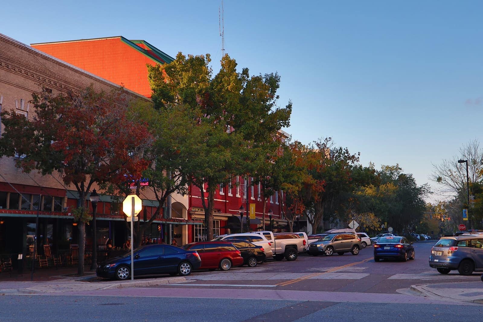 Downtown street corner with red-brick storefronts, autumn-colored trees and parked cars in warm evening light.