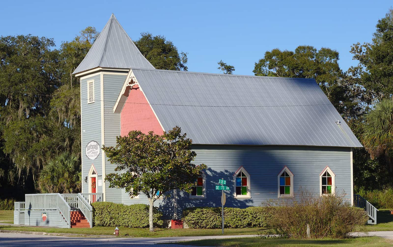 Small pastel blue and pink church-style building with a steeple and a metal roof under trees.