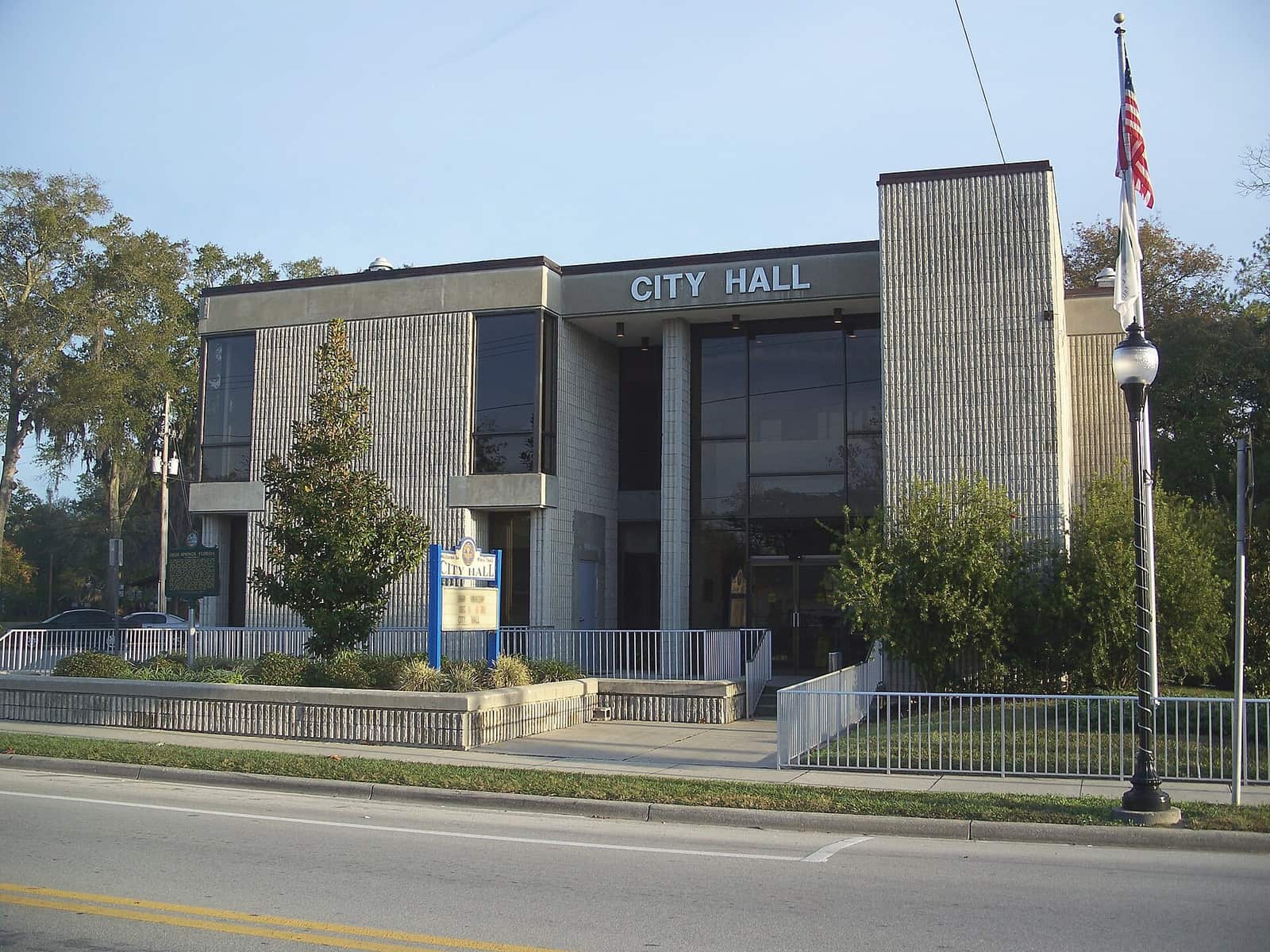 Modern two-story city hall building with a flagpole, a stone sign, and a flanking lawn beside the road.