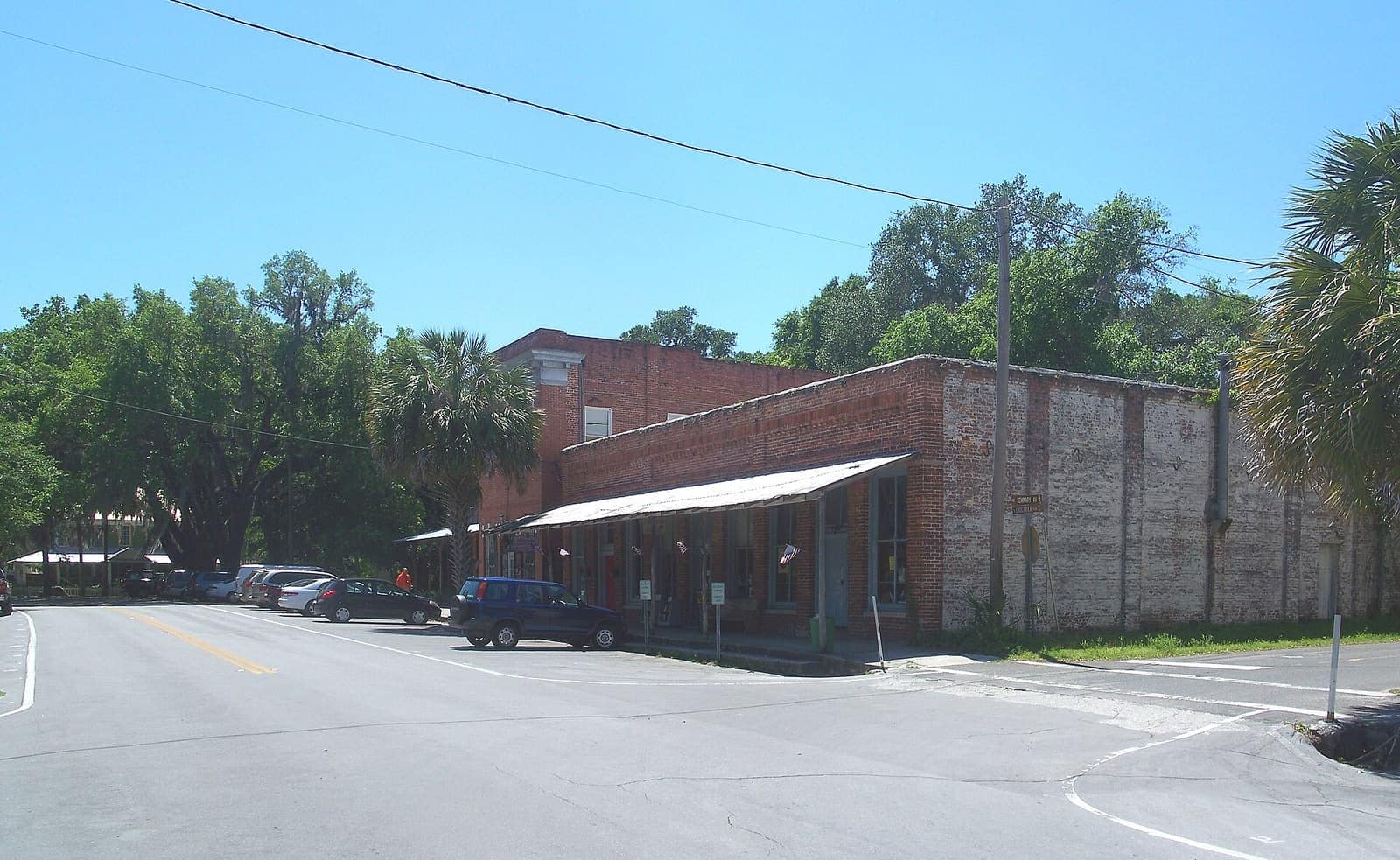 Brick commercial buildings with a striped awning along a street corner, parked cars, and tall shade trees under blue sky.