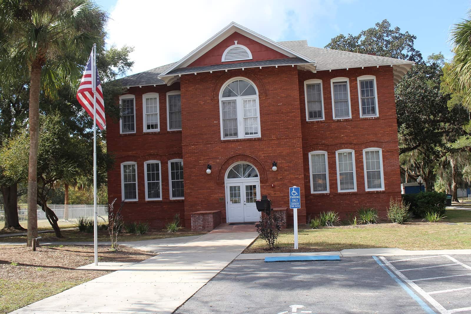 Two-storey red brick schoolhouse with arched windows and an American flag beside a paved walkway.