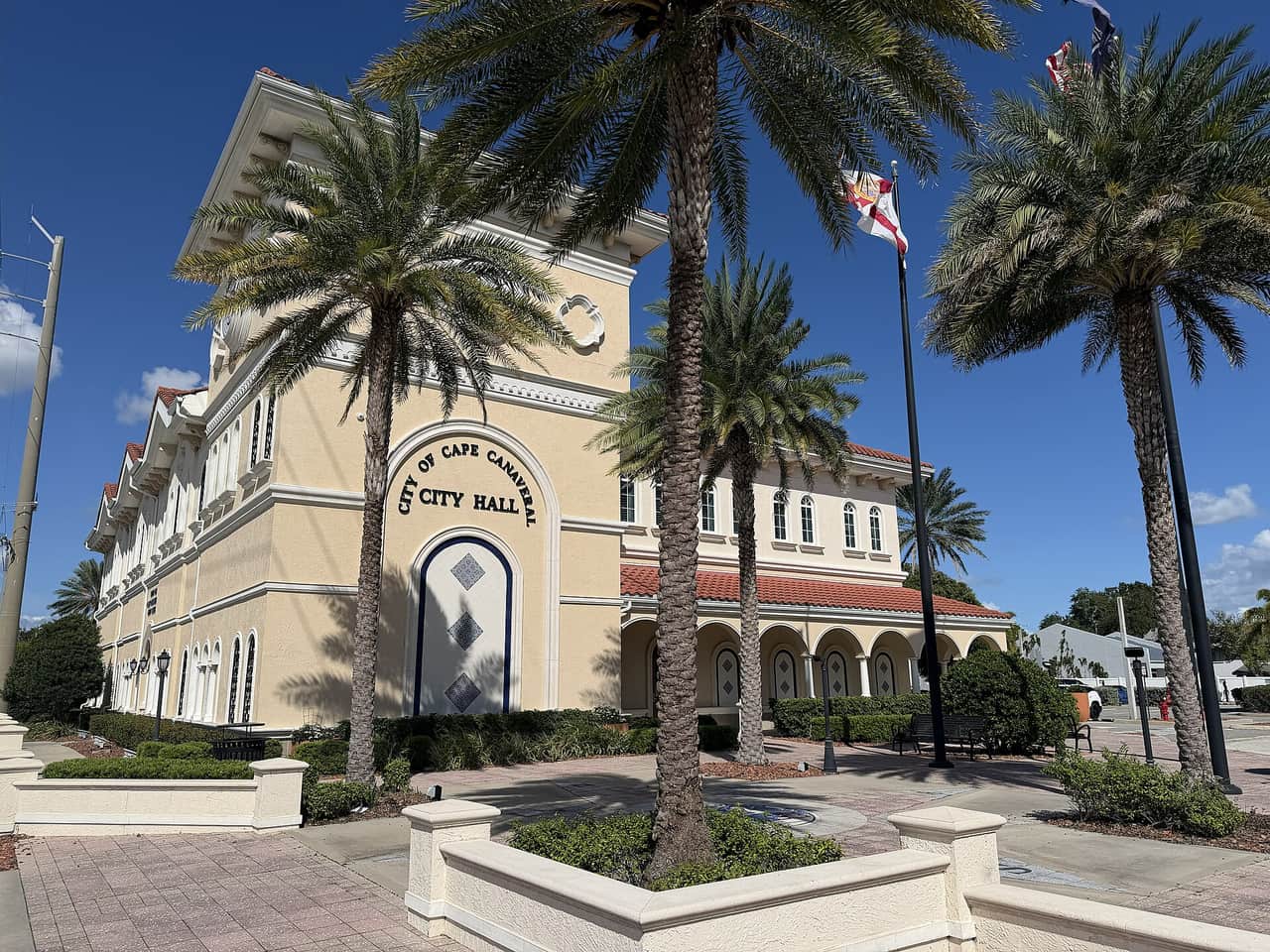 Cream stucco city hall building with arched entrance, palm trees, and flags under a bright blue sky.