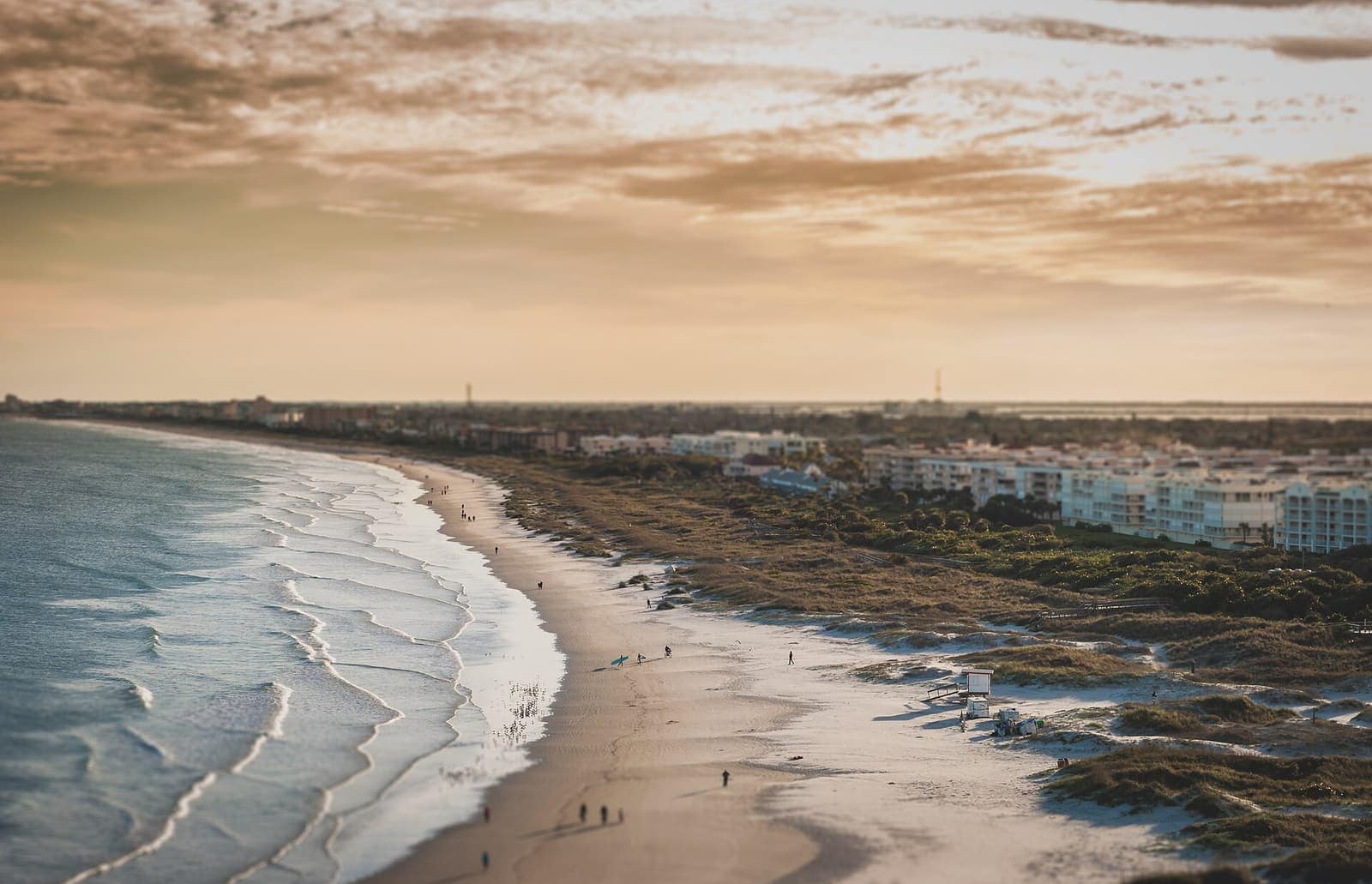 Aerial view of a sandy beach at dusk with waves, walkers on the sand, and a line of low buildings inland.
