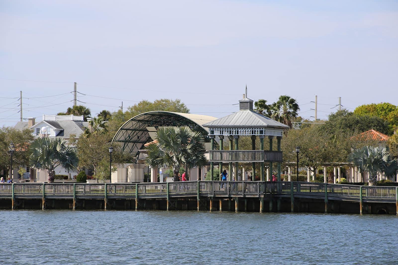 Covered pavilion and wooden boardwalk along a riverfront with palm trees and a few small distant people.