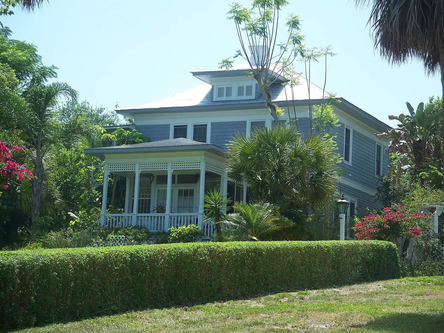 Two-story blue historic house with a white front porch, surrounded by hedges, palms and flowering shrubs.