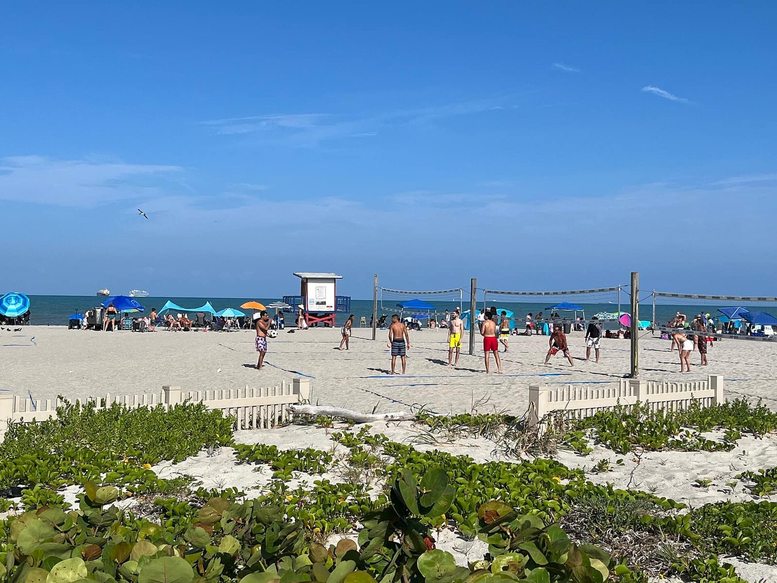 People play beach volleyball on sand near a lifeguard stand, with umbrellas and a dune fence in the foreground.