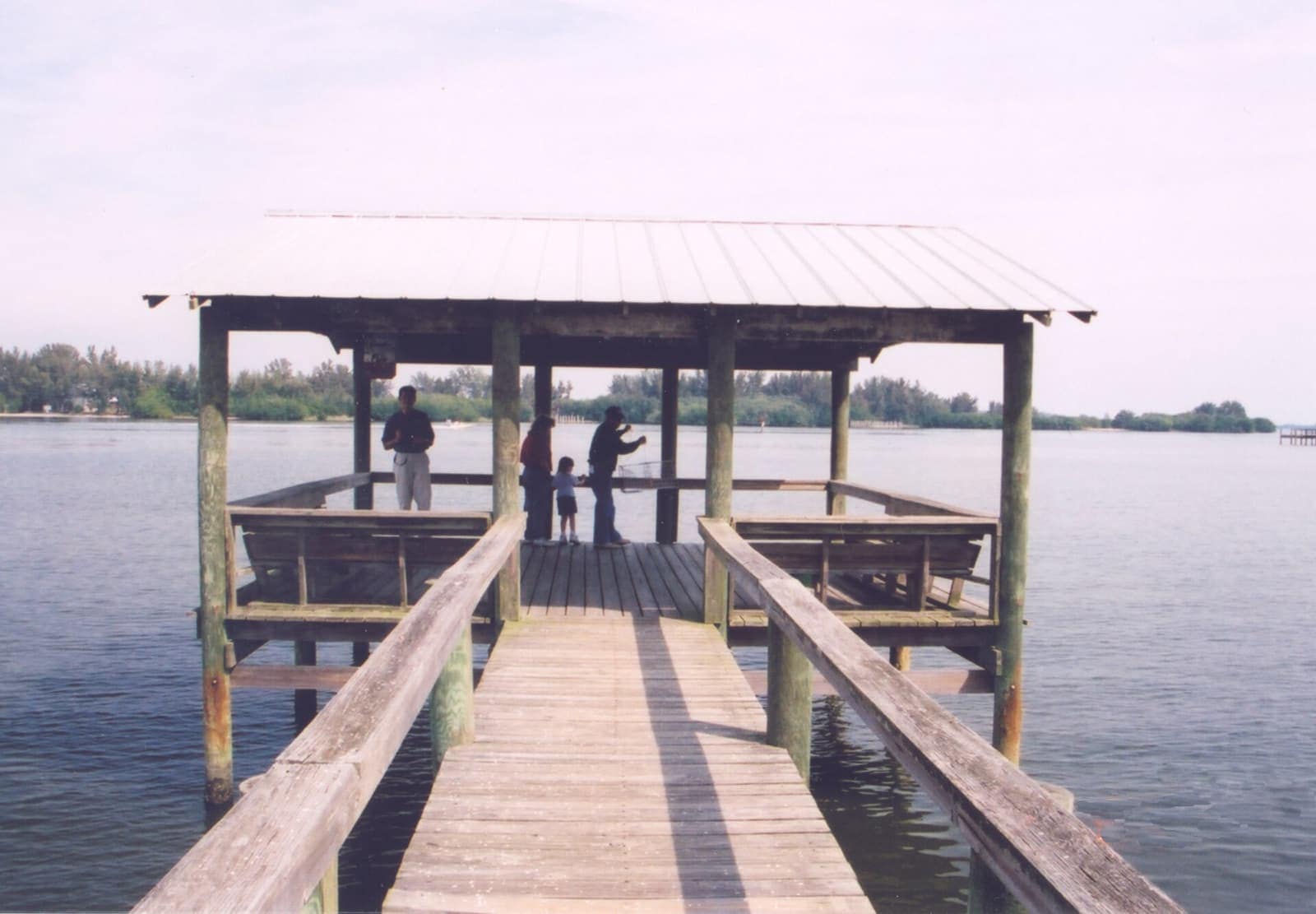 A covered wooden fishing pier extends over calm water, with a few small distant visitors standing near its end.