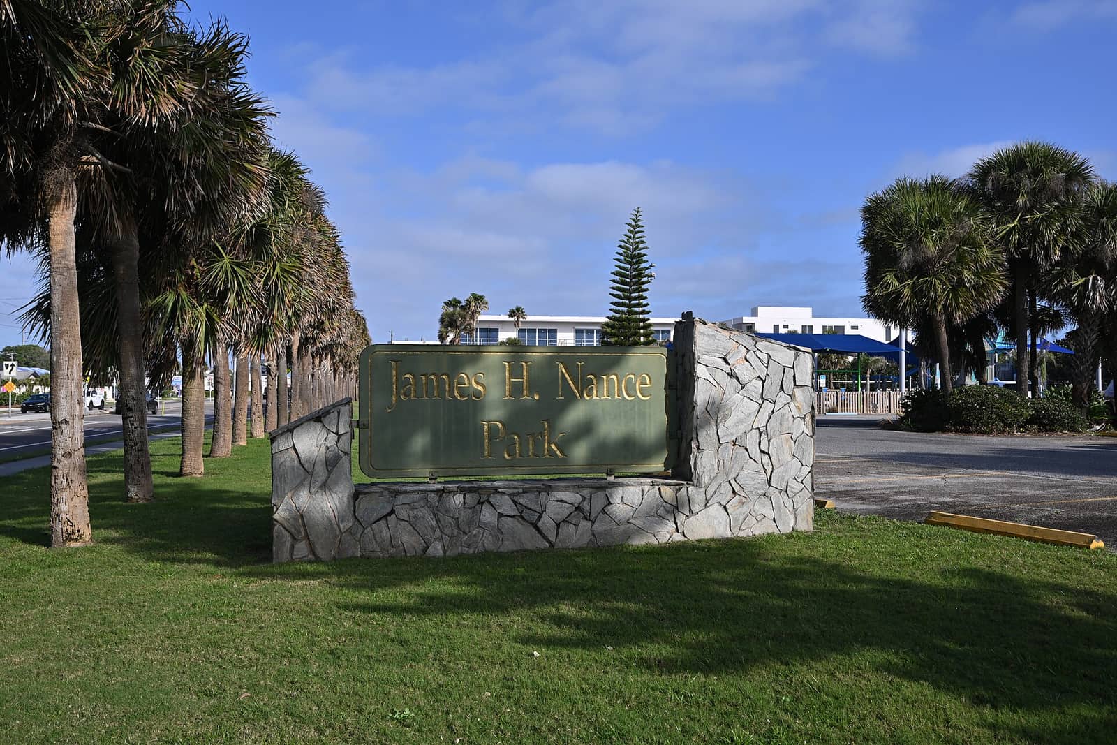 Green park sign reading James H. Nance Park on a stone base beside tall palm trees and a lawn.