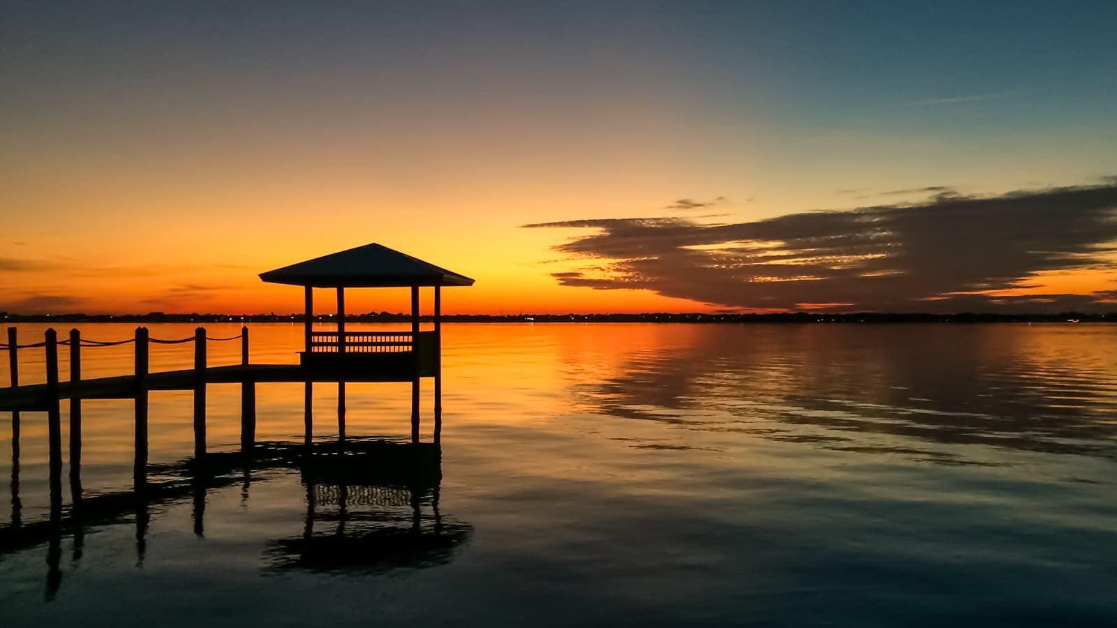 Silhouetted wooden gazebo on a dock over calm water at sunset.