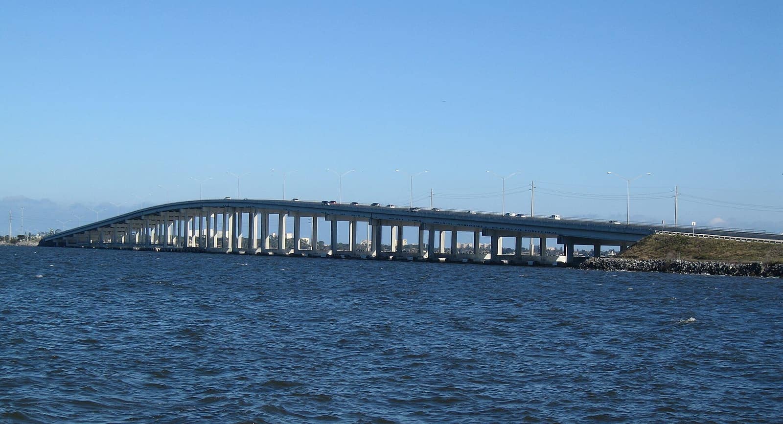 Long multi-span concrete causeway bridge crossing blue water under clear sky.
