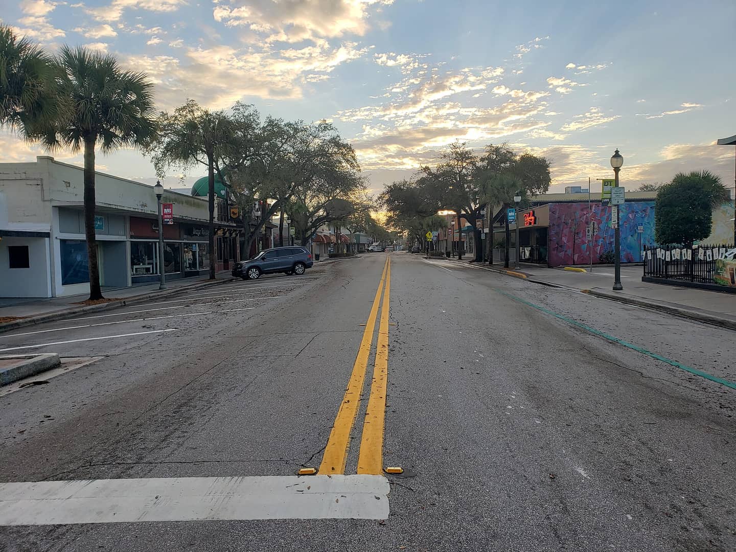 Empty downtown street with yellow center lines, palm trees and storefronts under a dusk sky.