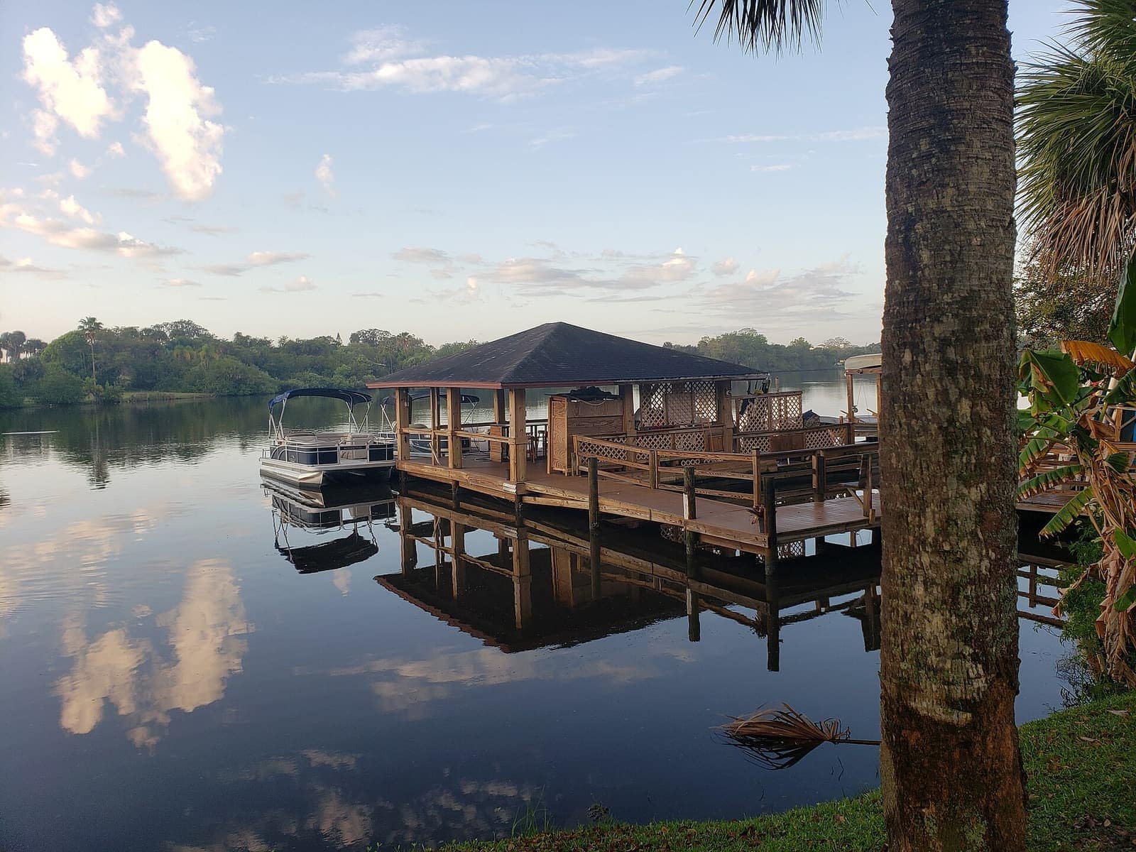 A covered wooden dock extends over calm water with a pontoon boat moored beside it.