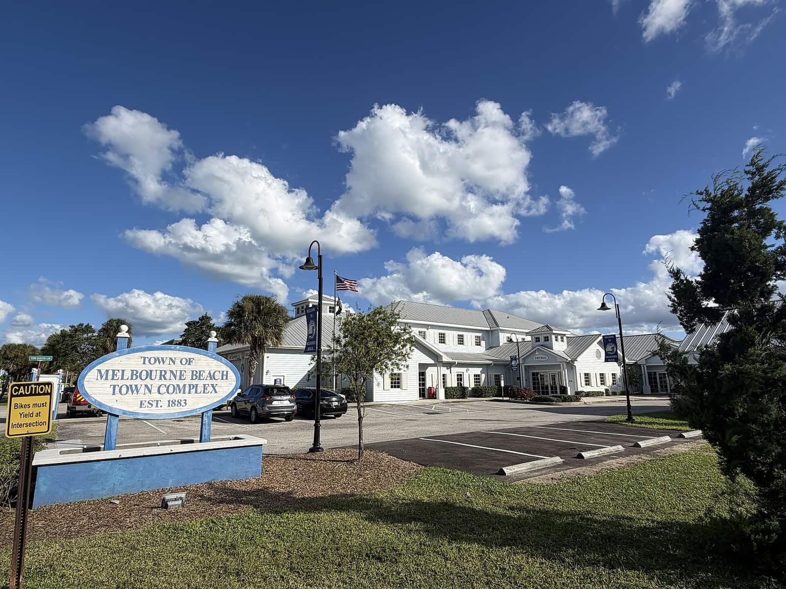 White two-story town complex building behind a blue oval sign and parked cars under cumulus clouds.