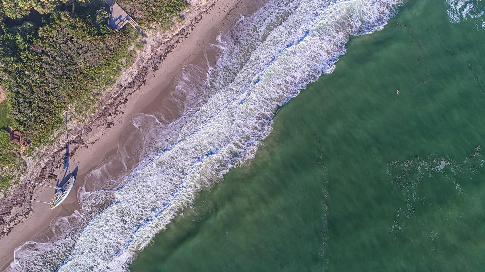 Aerial view of sandy beach, breaking white surf, green water and a beached sailboat on shore.