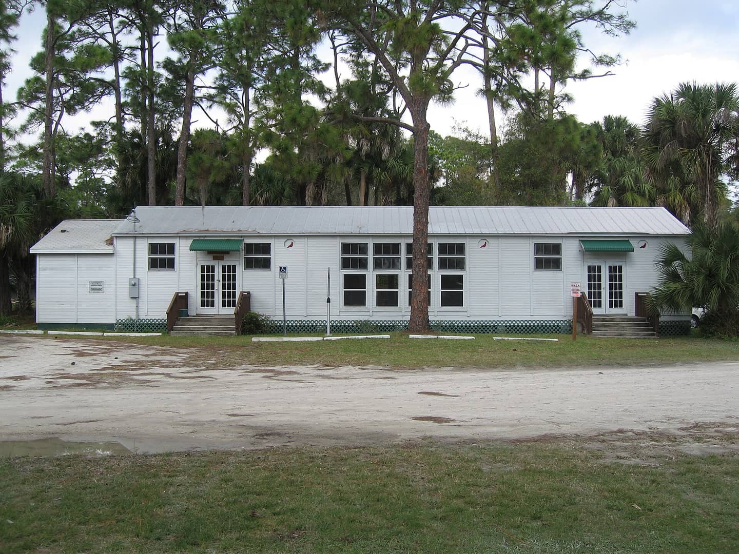 White wooden community hall with green awnings and pine trees, under a pale sky.