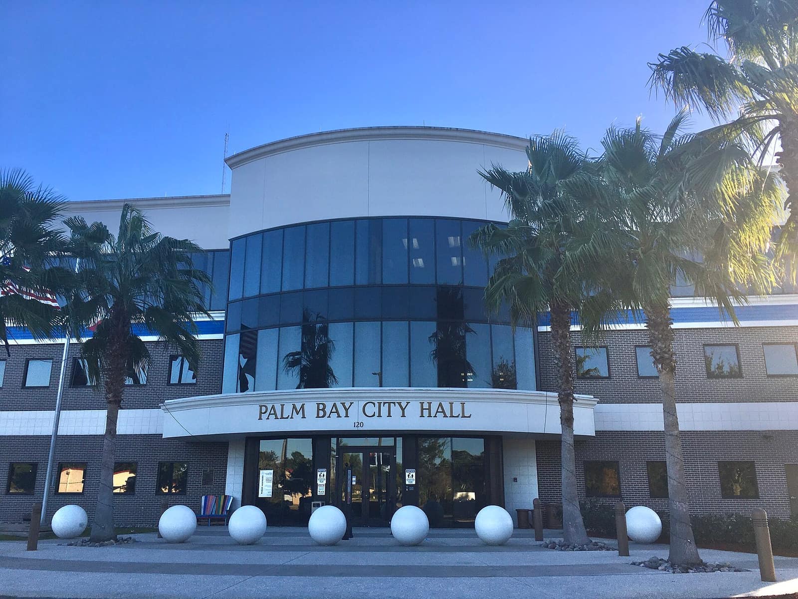 Front entrance of a two-story brick municipal building with palm trees and round white planters under blue sky.