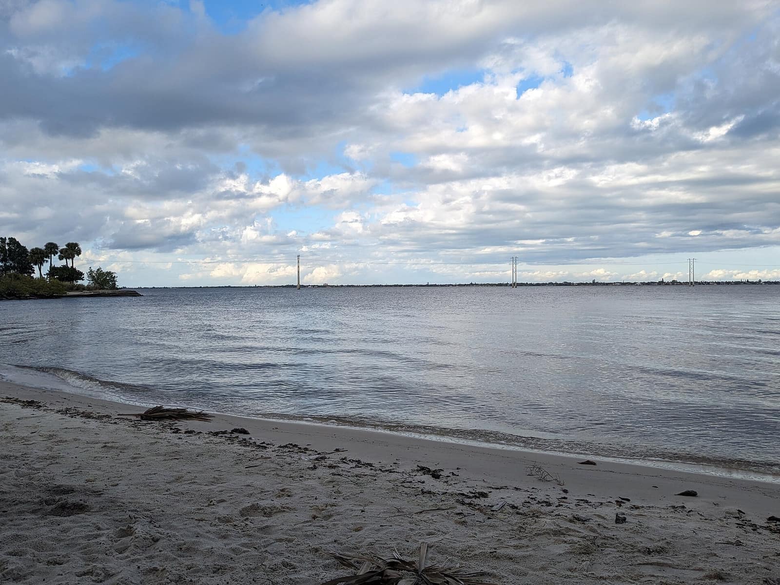 Wide view across a calm bay from a sandy beach under a cloudy sky, with distant towers on the far shore.