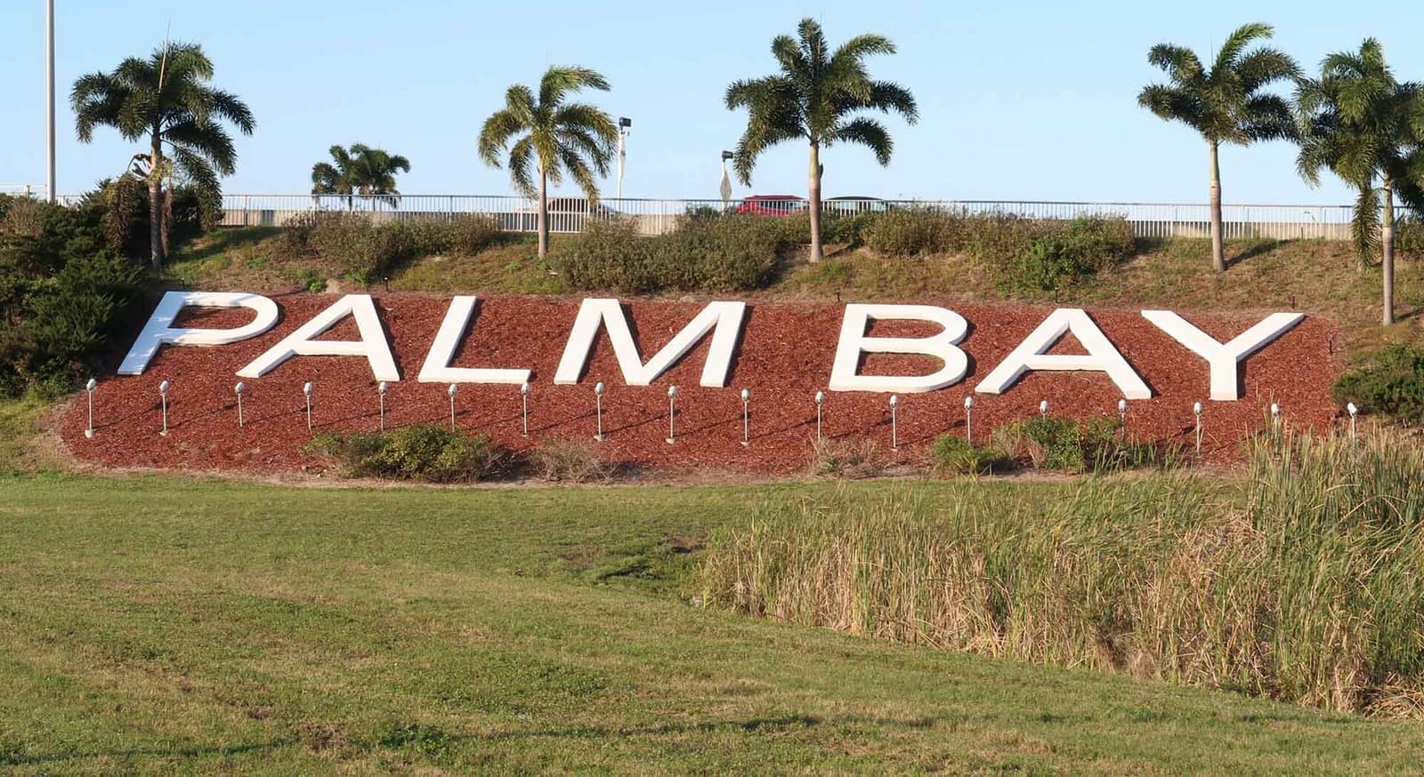 Large white PALM BAY letters on a mulch bed beside a grassy slope with palm trees behind.
