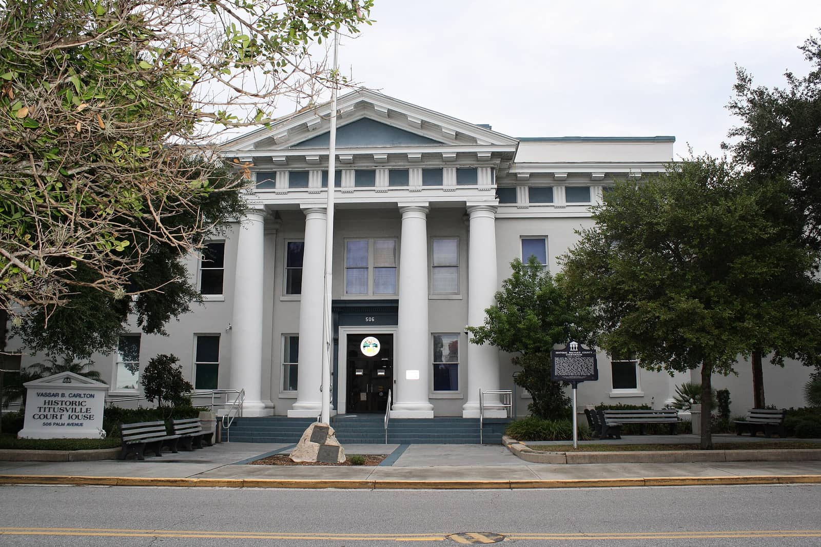 White columned historic courthouse with a flagpole, benches, and trees under a pale sky.