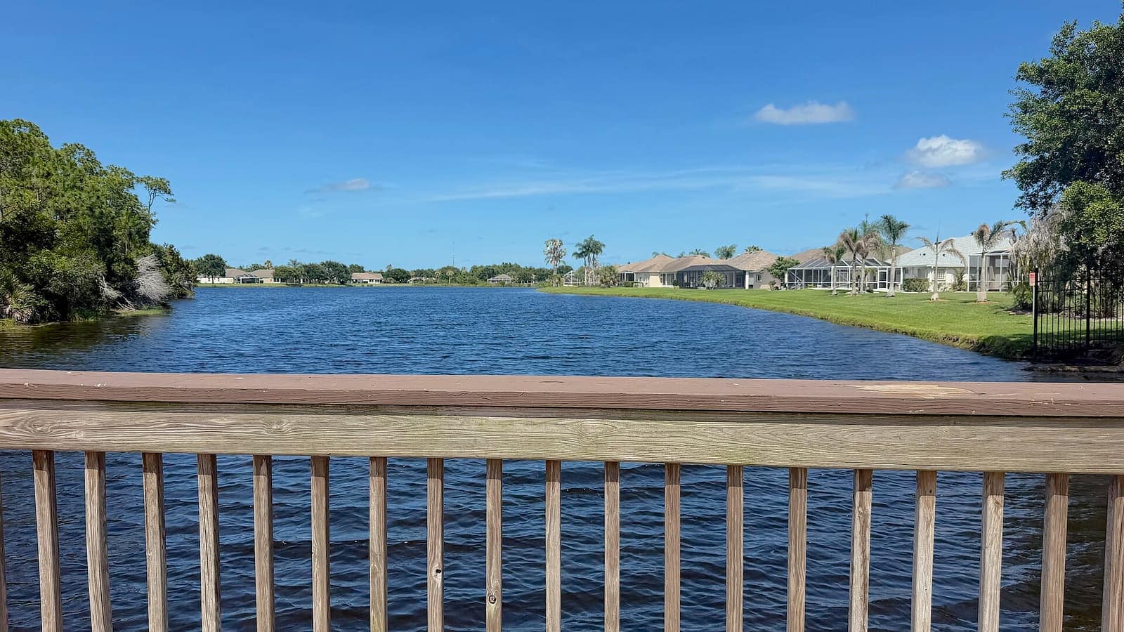 A calm lake viewed past a wooden railing, with waterfront homes and palm trees under a blue sky.