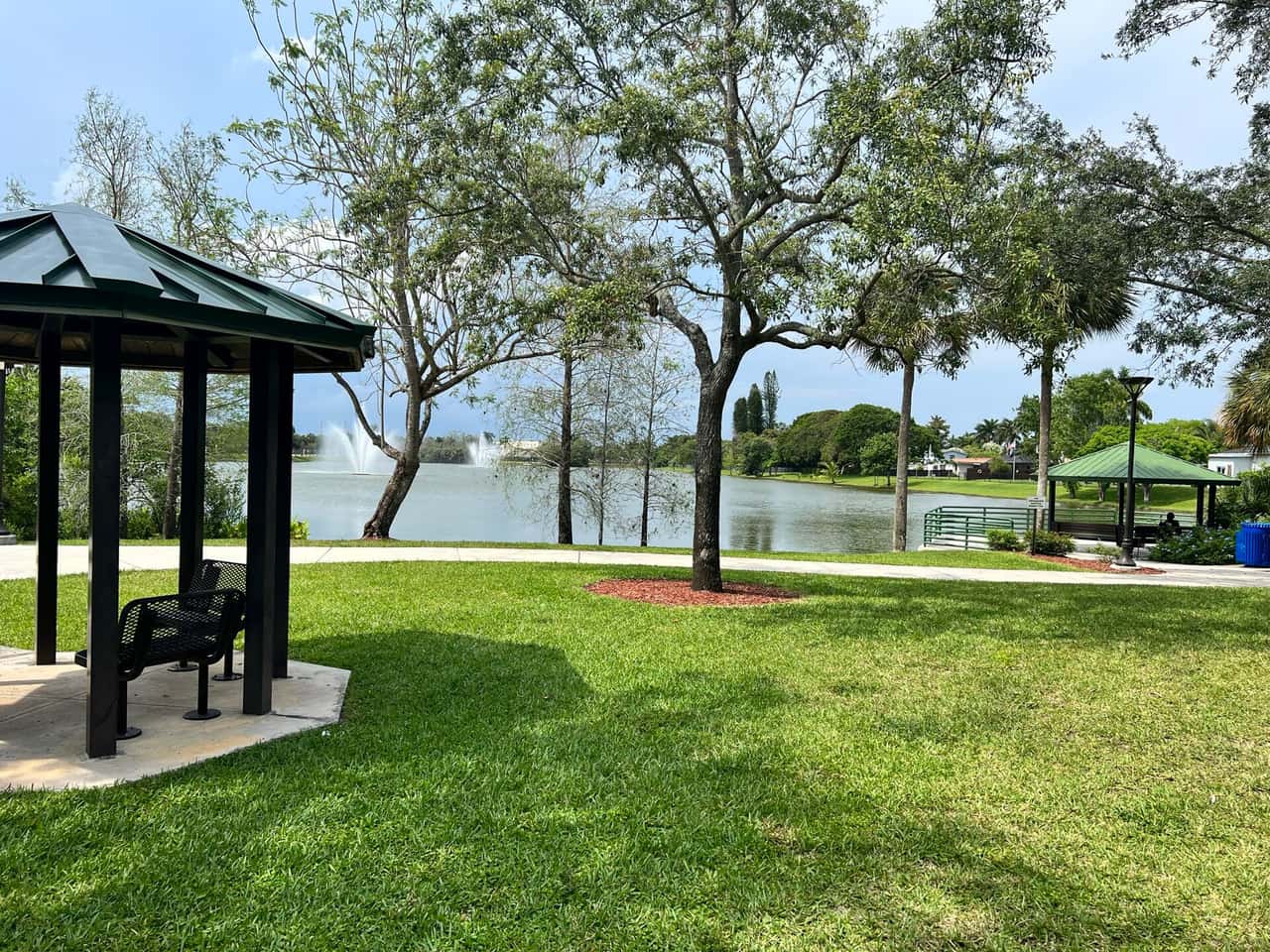 Covered gazebo overlooks a lake with fountain and grassy park lawn under trees.