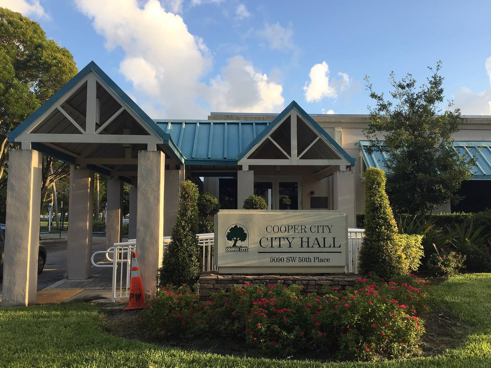 Cooper City City Hall entrance with a teal-roofed covered walkway, a sign, and flowering shrubs.