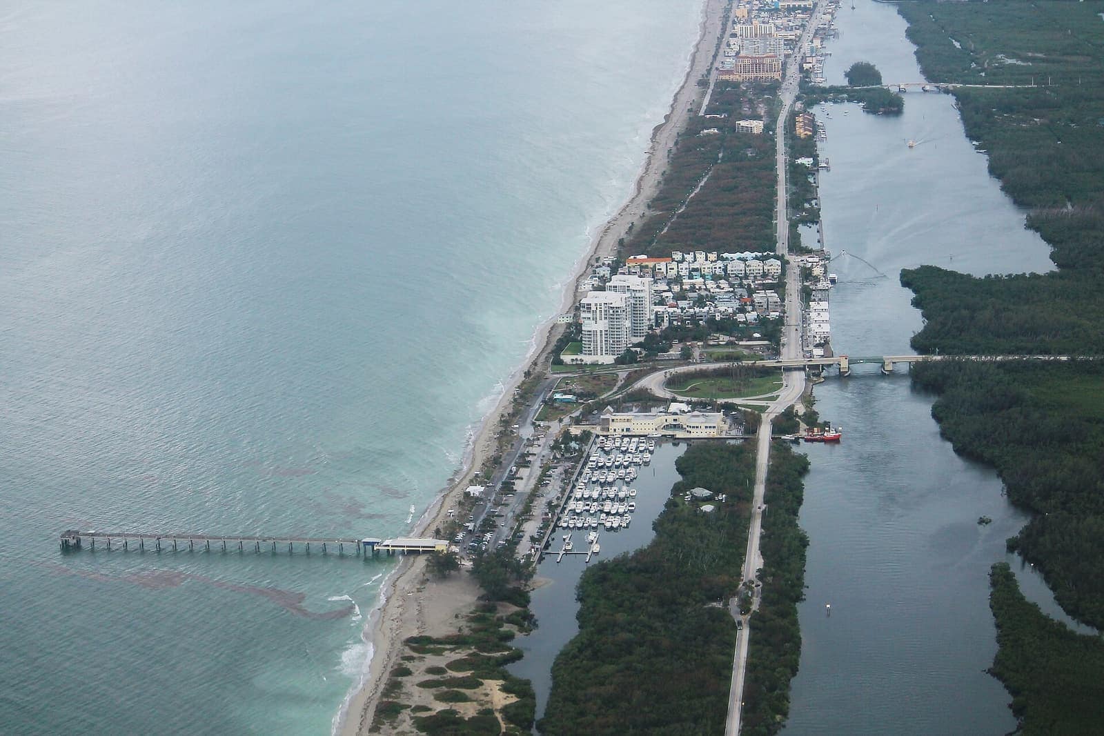 Aerial view of a thin coastal strip with a long fishing pier, marina boats, high-rise buildings and mangrove waterways.