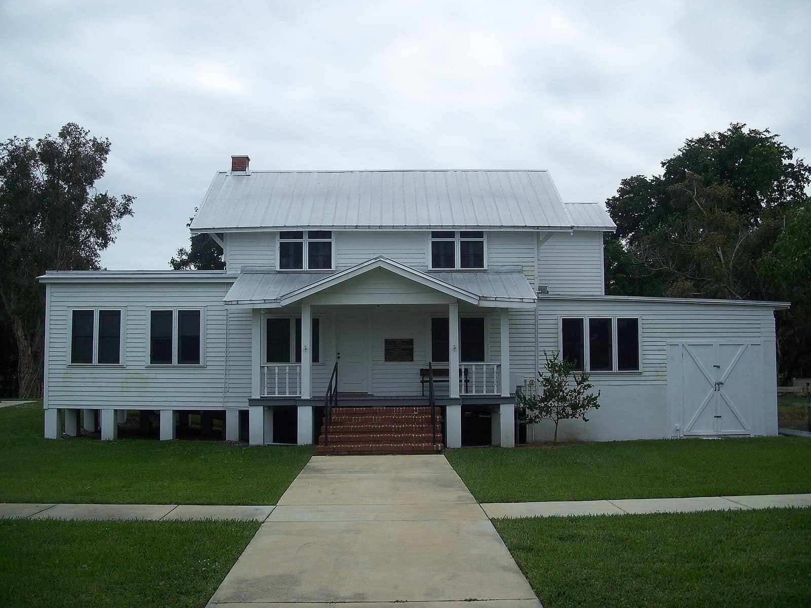 Two-story white wooden house with a metal roof, front porch steps, and a lawn under overcast sky.