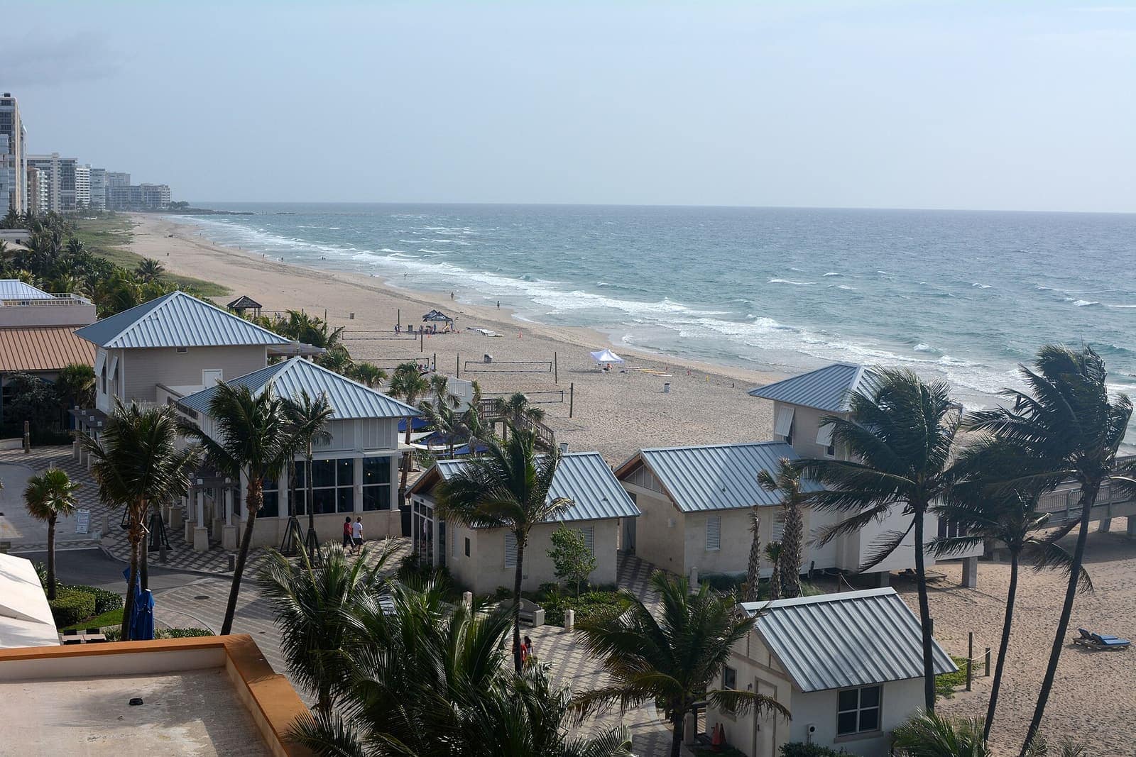 Aerial view of a sandy beach, ocean waves, low cabanas with metal roofs, and palm trees.