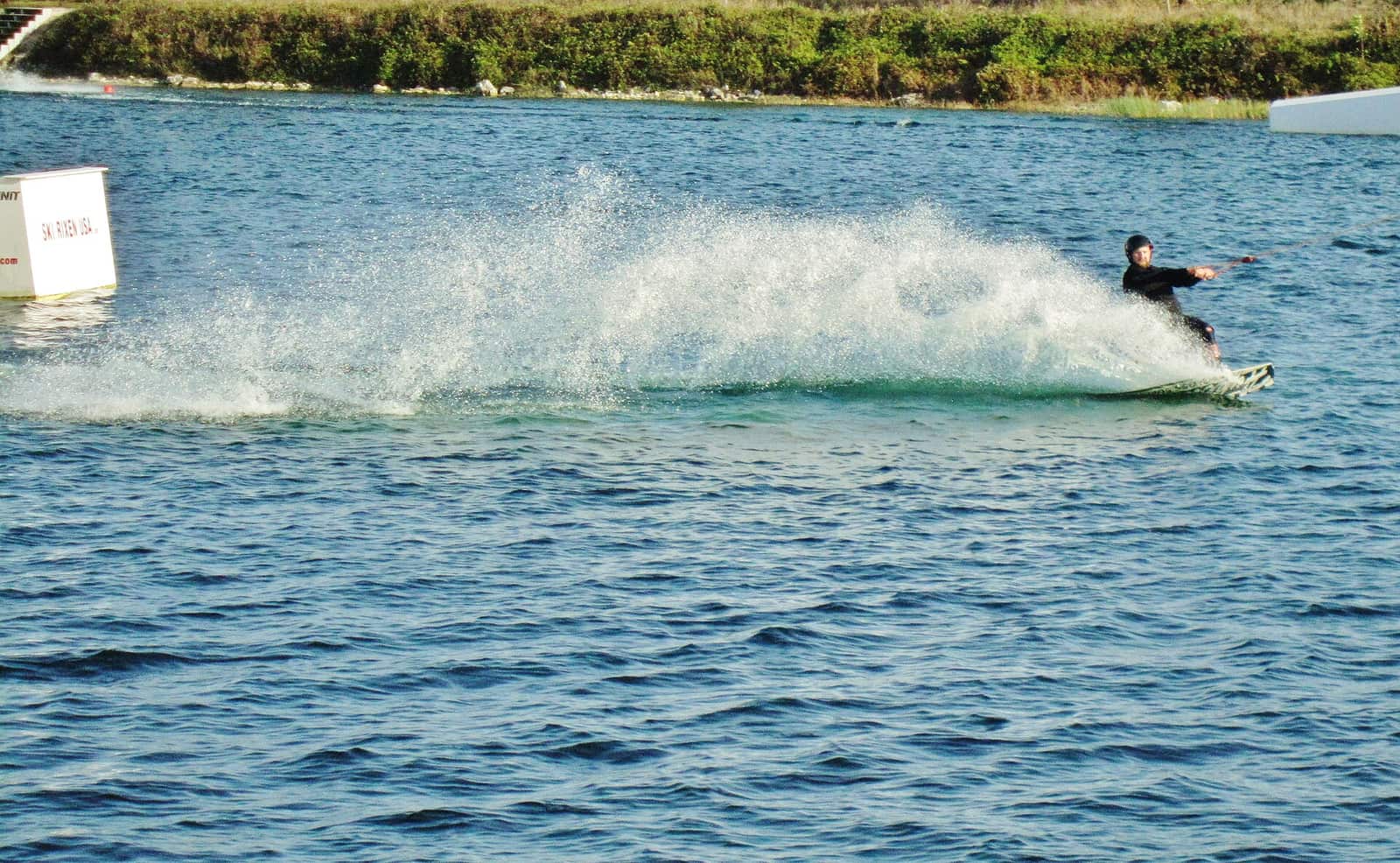 A cable water skier sprays a wake across a blue lake near a white buoy and shoreline.