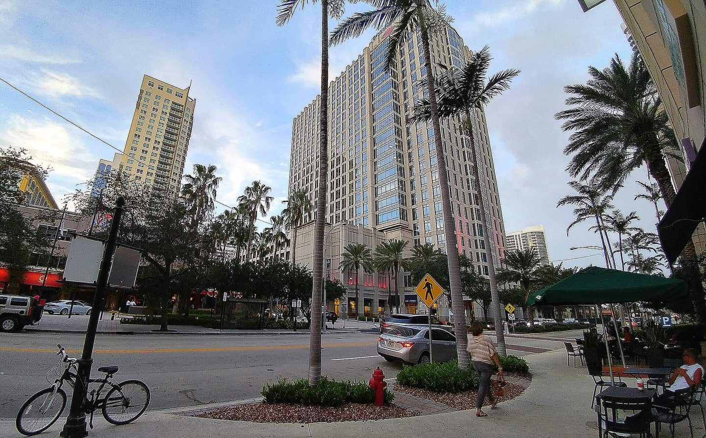 Downtown street scene with a tall hotel tower, palm trees, a parked car, and a bicycle at the curb.