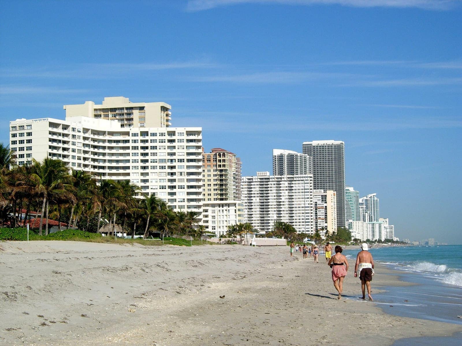 Sandy beach with walkers near the surf, and a row of high-rise condominium towers under blue sky.