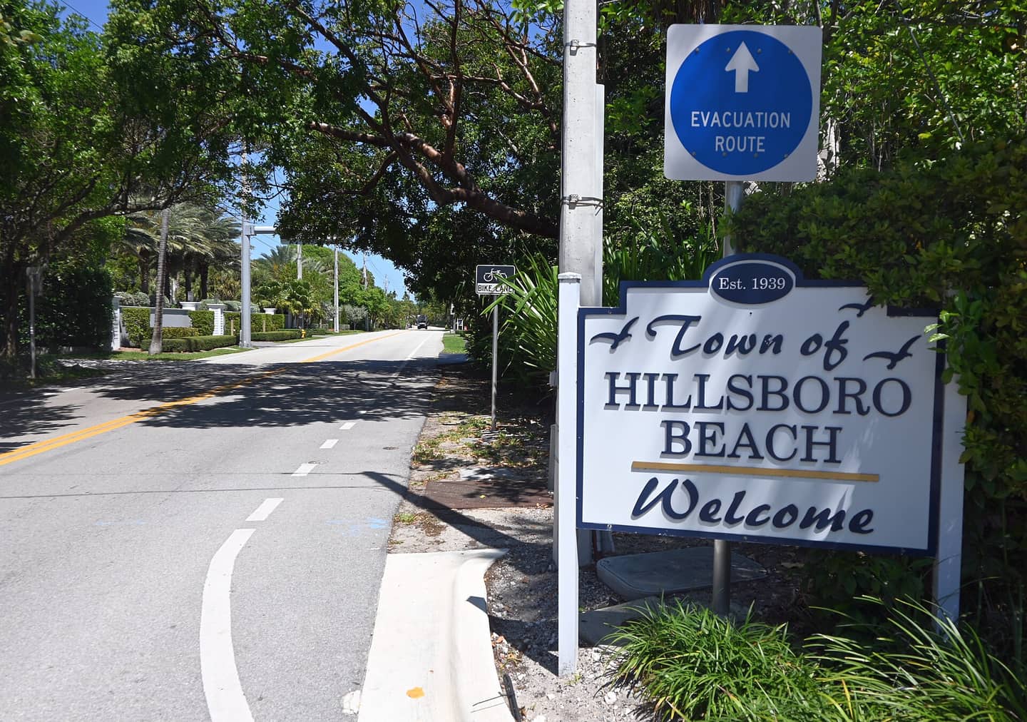Town of Hillsboro Beach welcome sign beside a tree-lined road and an evacuation route sign.