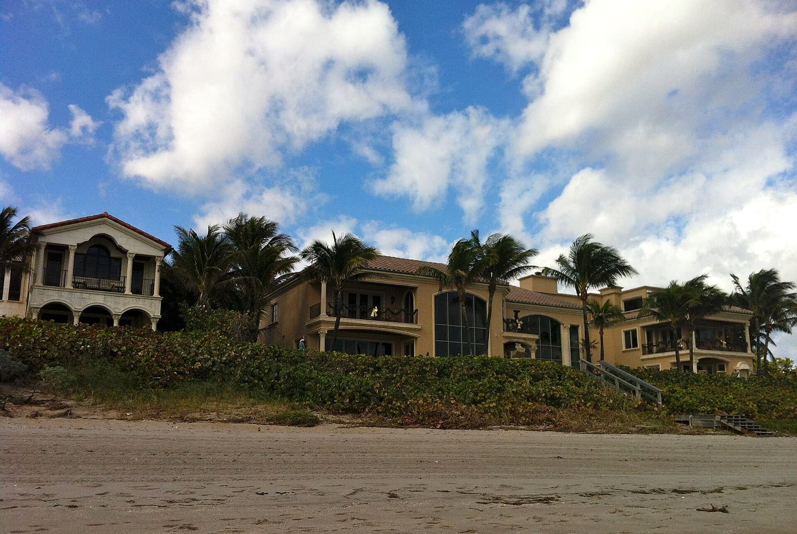 Large beachfront homes with balconies and palm trees rise above a sandy beach under a cloudy blue sky.