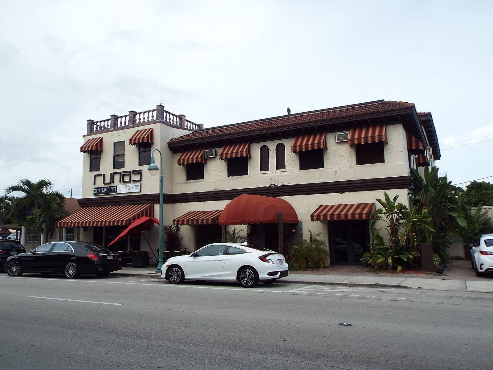 Two-story Mediterranean-style building with striped awnings, a tiled roof, and palms along a quiet street.