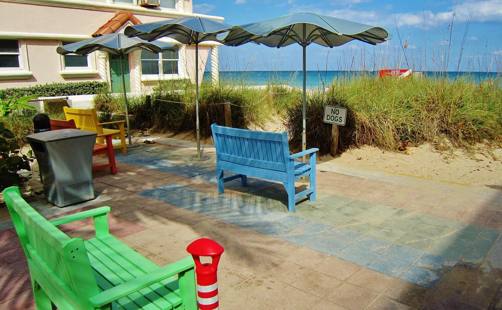 Colorful benches and grey umbrellas on a paved patio beside sea oats and the ocean.