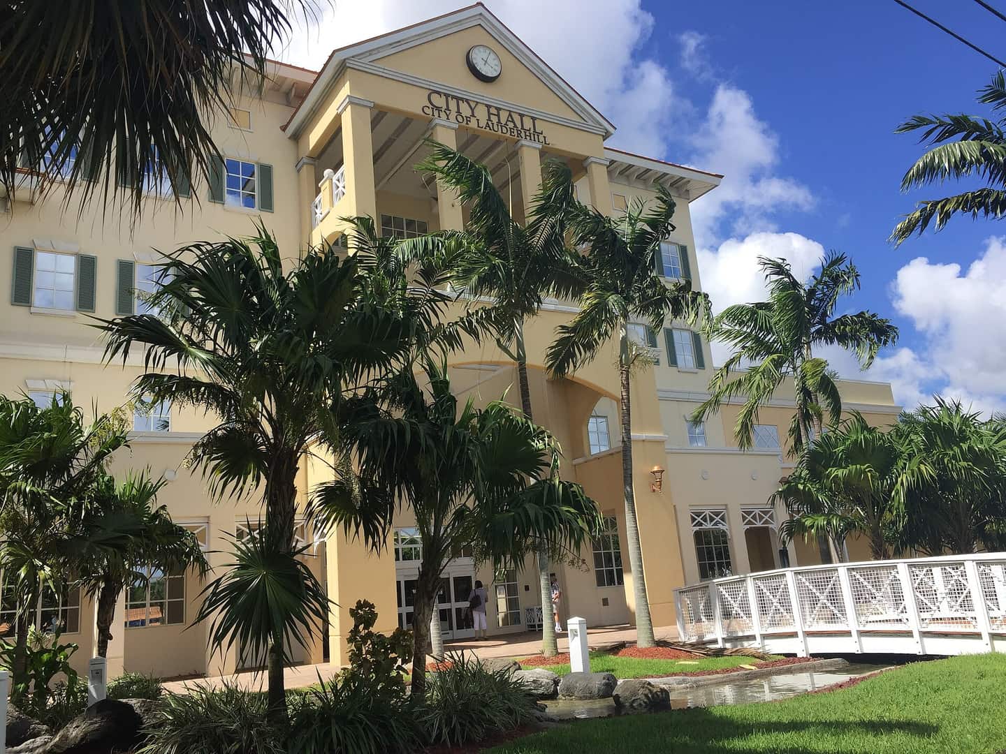 Cream three-story city hall building with columns, palm trees, and a white footbridge under blue sky.