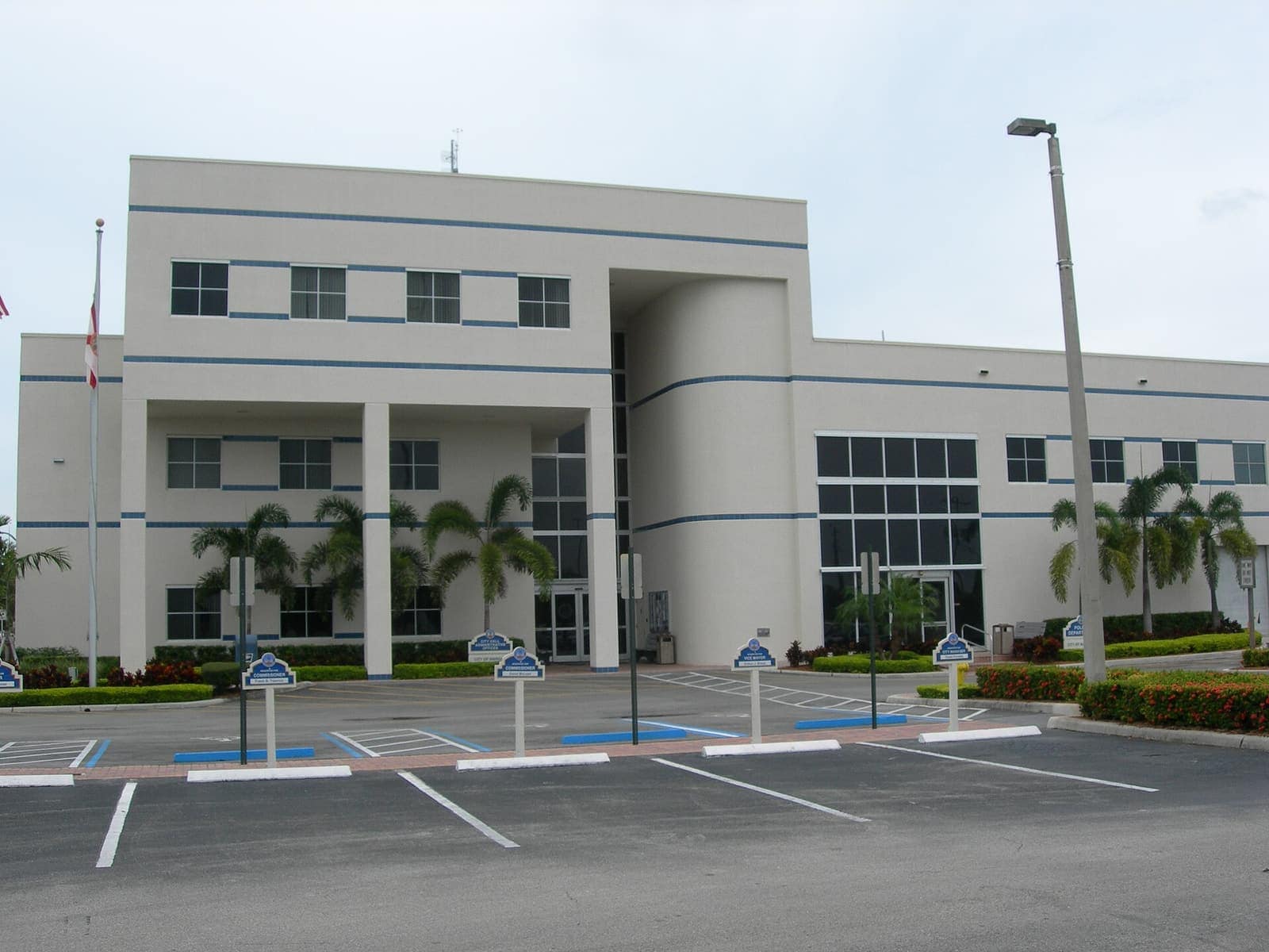 Three-story cream stucco municipal building with blue stripes, palm trees, and a nearly empty parking lot under overcast sky.
