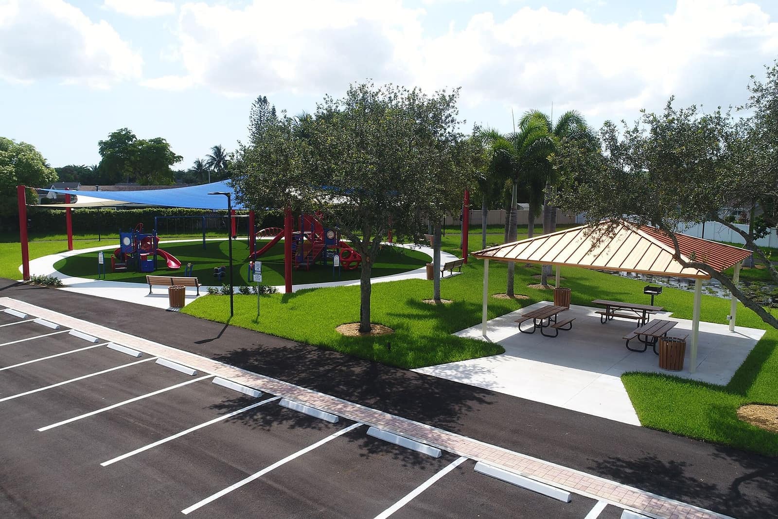 Green park with a red playground structure, shaded picnic tables under a wooden pavilion, and a paved parking lane.