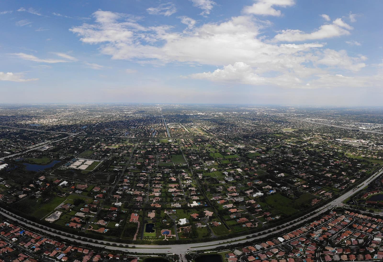 Aerial view over a dense grid of suburban homes, roads and lakes under partly cloudy sky.
