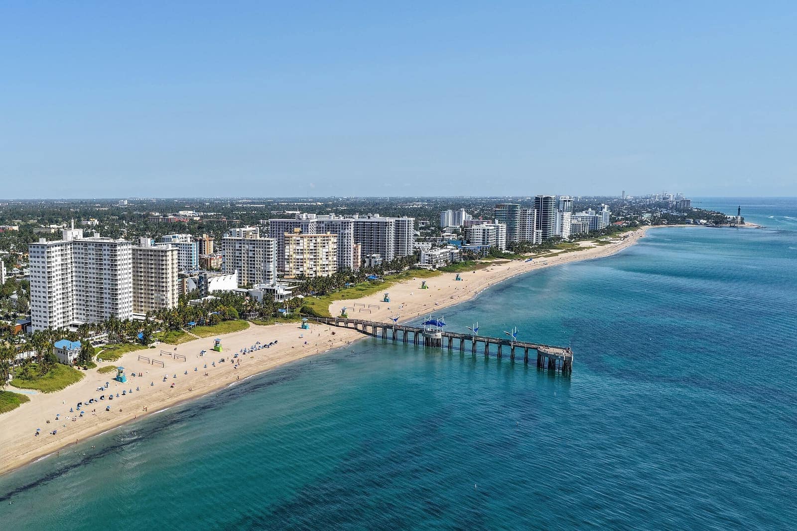 Aerial view of a sandy beach, high-rise towers, and a long fishing pier over turquoise water.
