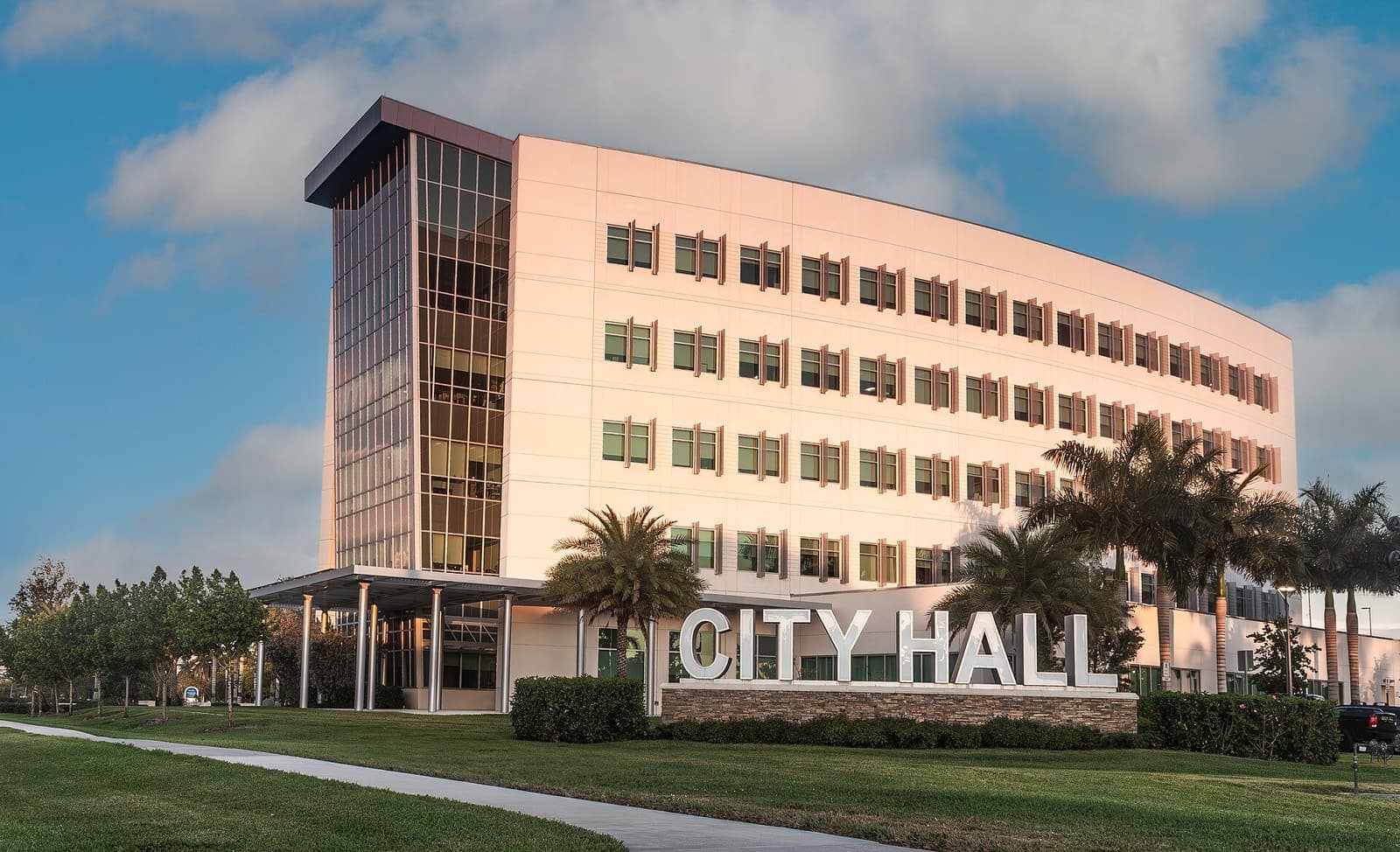 Curved multi-storey city hall building with large CITY HALL letters on a stone wall and palm trees in front.