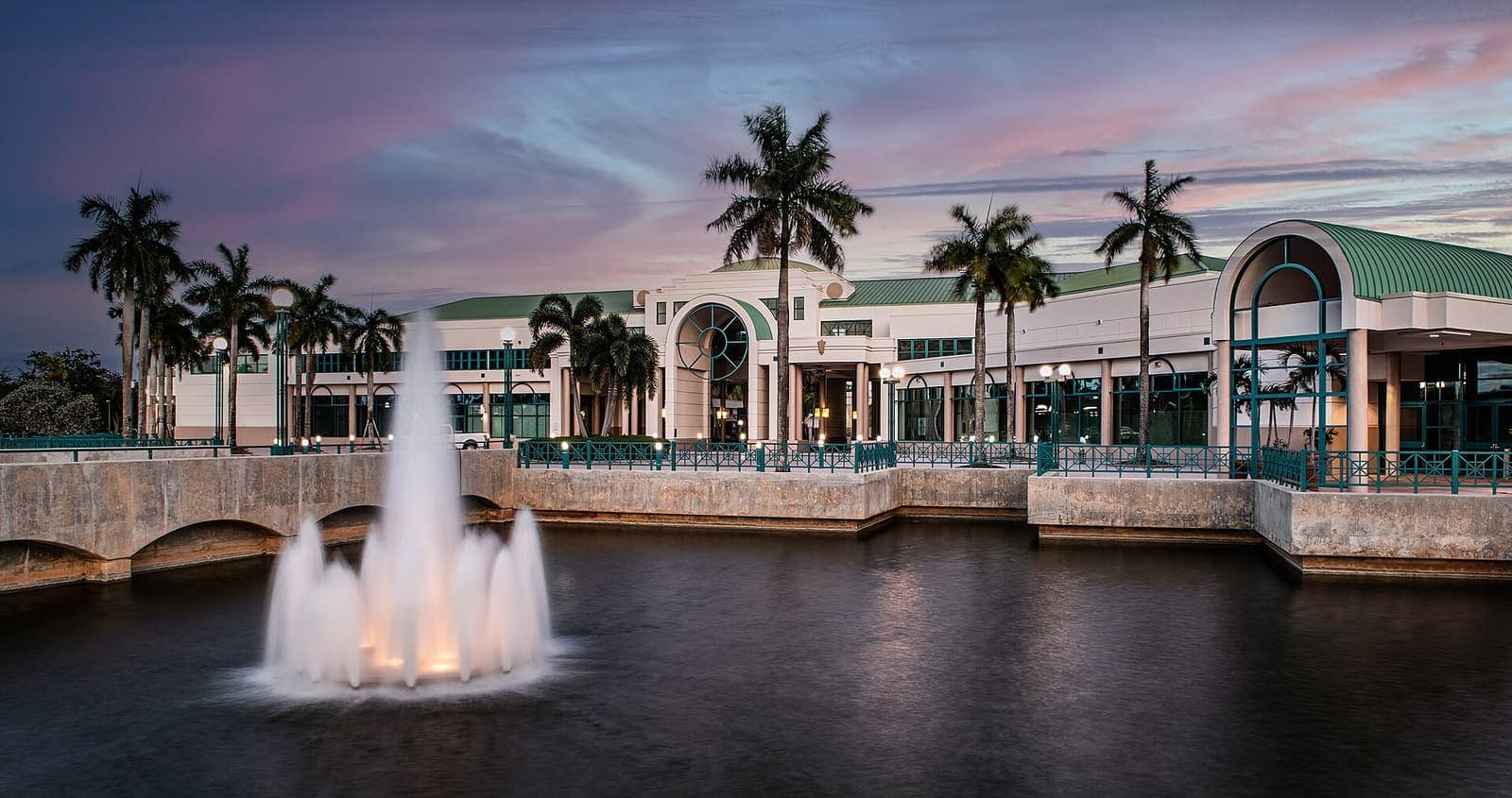 Modern civic building with arched entrance and palms beside a fountain spraying over a dusk-lit pond.