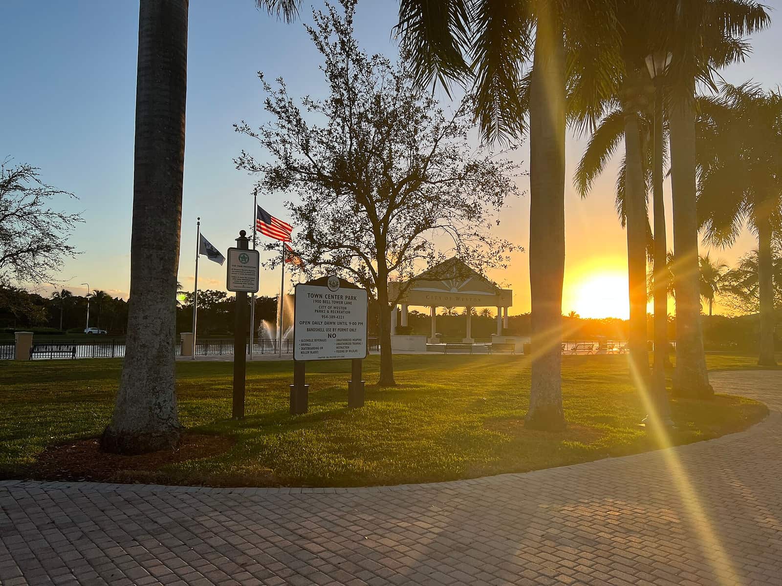 Sunset over a Florida park with flags, a gazebo, palm trees and a paved walkway.