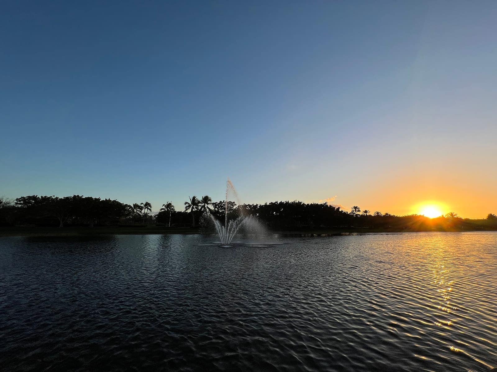 Wide view of a lake fountain spraying at sunset with rippling water and a clear sky.