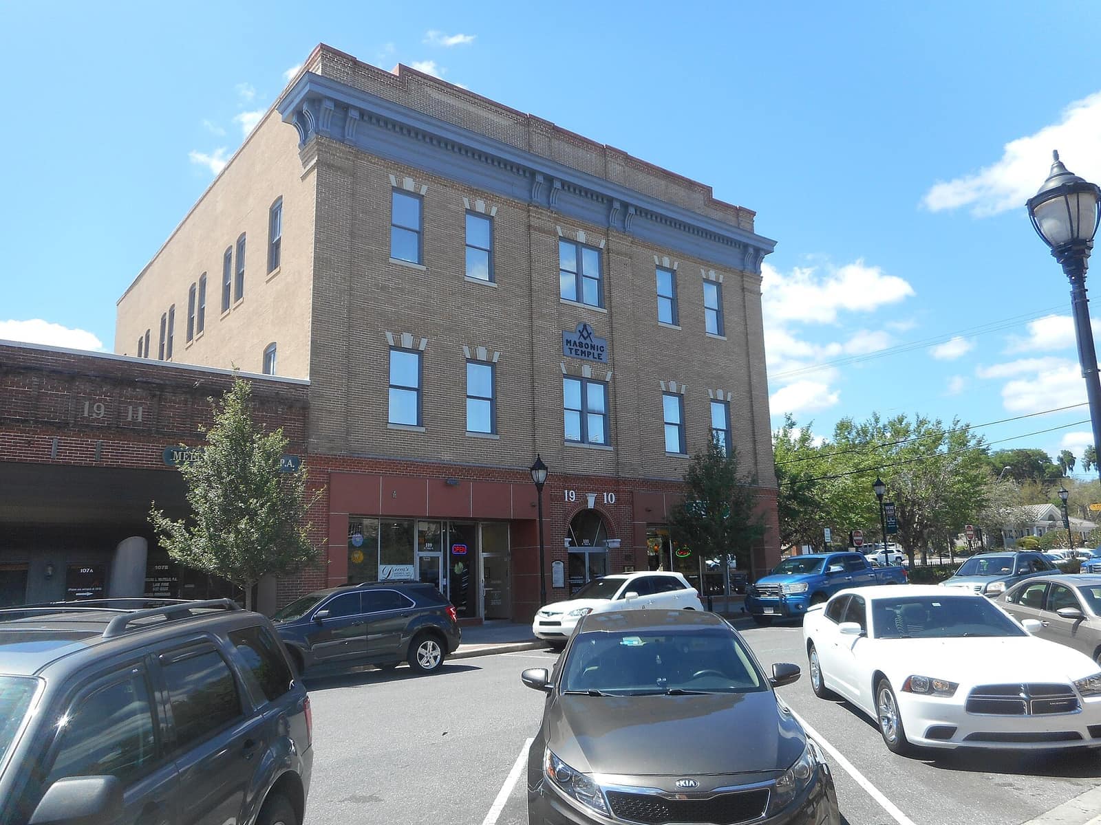 Three-story tan brick historic building with a blue emblem and parked cars in the foreground on a sunny day.