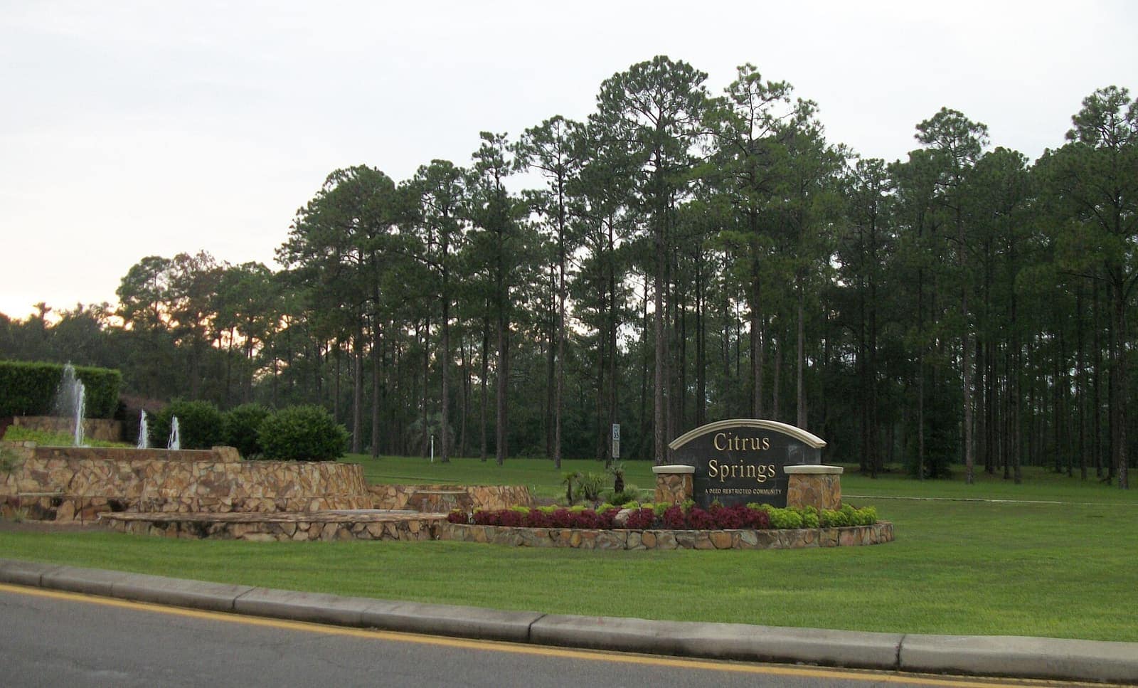 Green-and-gold Citrus Springs entrance monument with a stone fountain, flower beds, and tall pine trees behind a lawn.