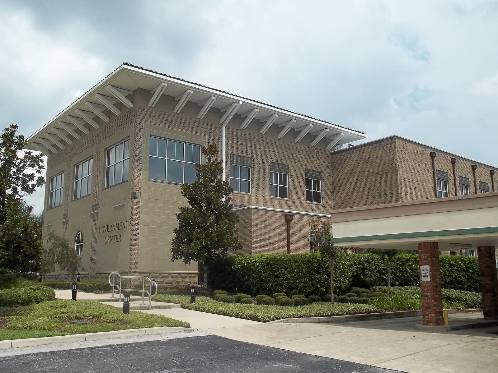 Two-story tan brick government center building with a covered drive-through entrance and landscaped shrubs.